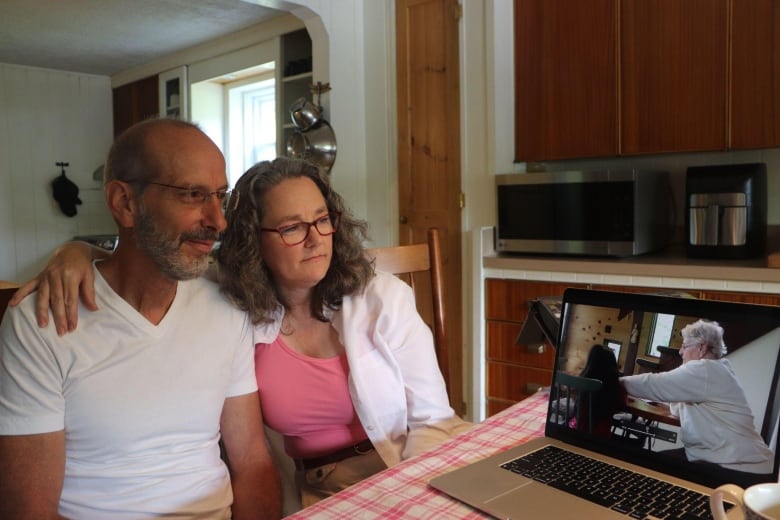 A couple sits at their kitchen table looking at a home video on their computer 