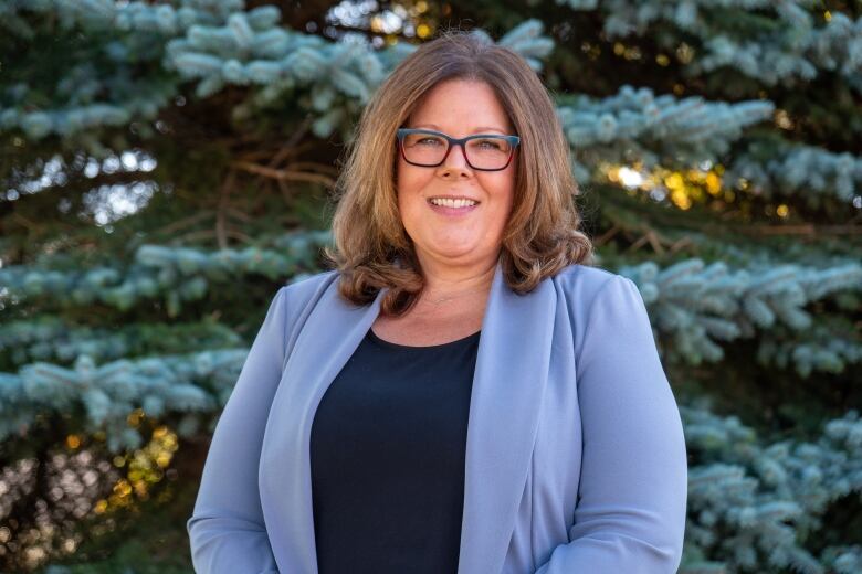 A person wearing glasses and a light blue blazer stands outside in front of some pine trees, smiling.