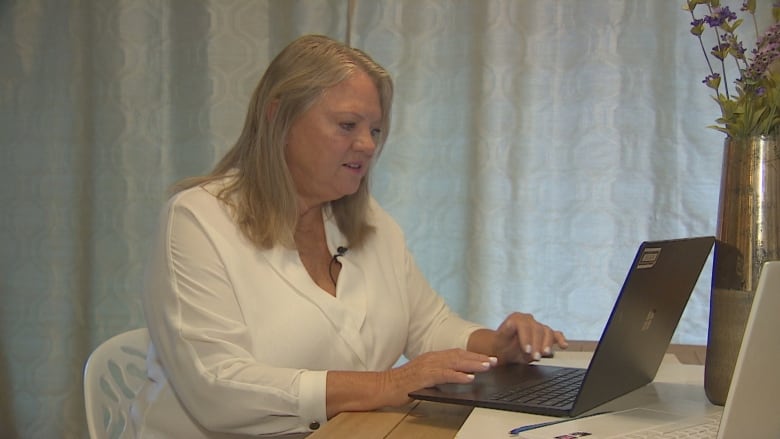 A blond-haired woman in a white top uses a laptop that is sitting on her desk.