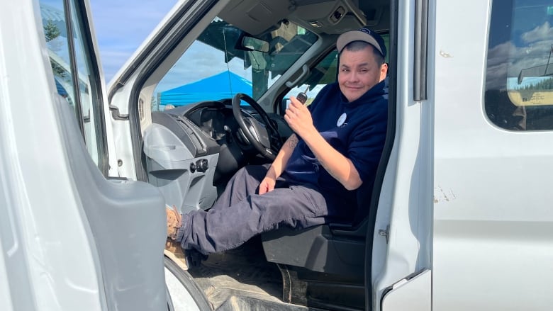 A man with a baseball cap and blue hoodie sits in a white transport van, holding up the keys.