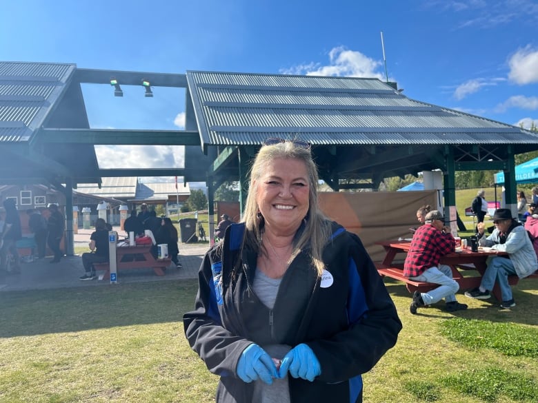 A women with a black and blue jacket and blue latex gloves smiles in front of a group of people.