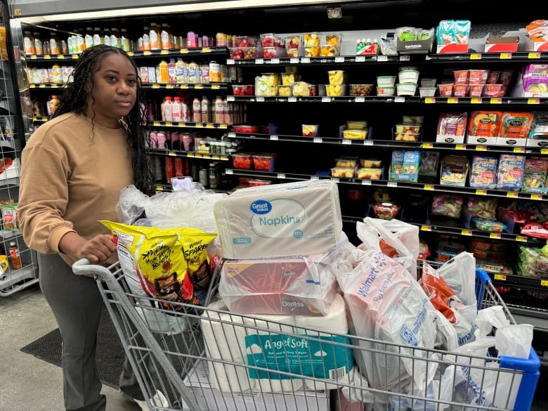 A woman pushes a shopping cart full of items in front of a refrigerated grocery aisle.