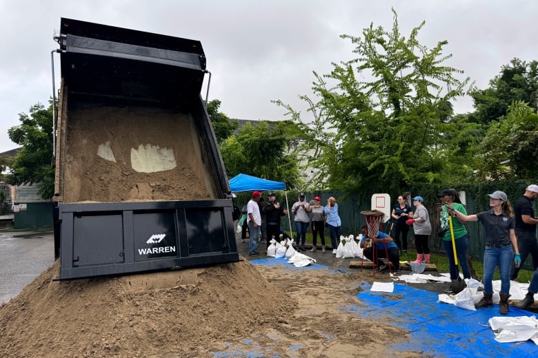 Sand pours from the back of a dump truck onto a blue tarp as people stand beside with shovels and white plastic bags. 