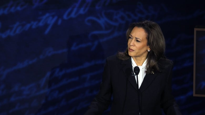 A woman in a dark suit stands behind a podium on a debate stage.