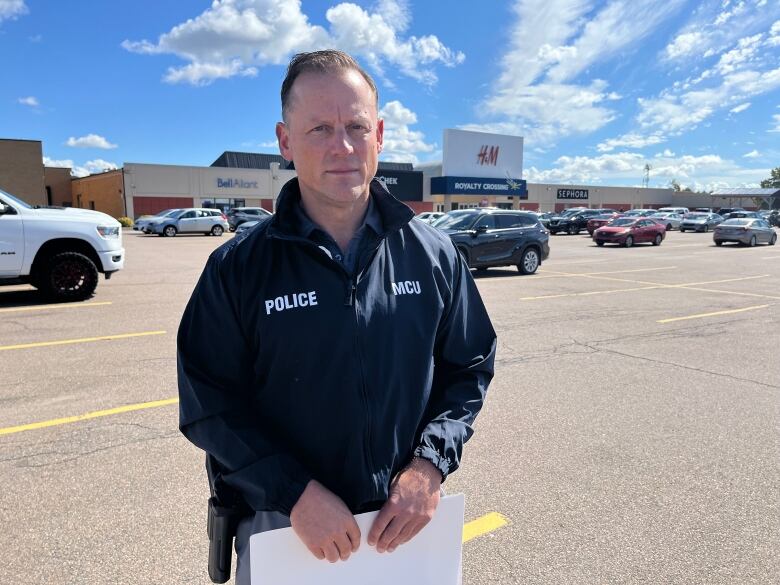 A man with short light hair wearing a blue jacket that says 'police' stands in a parking lot in front of a shopping mall.