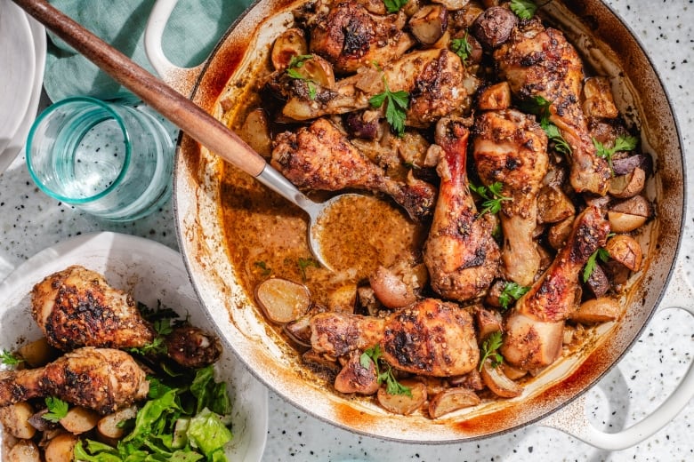 Overhead shot of a white pot with cooked chicken drumsticks and potatoes. A drinking glass and a bowl with the chicken sit next to the pot. 