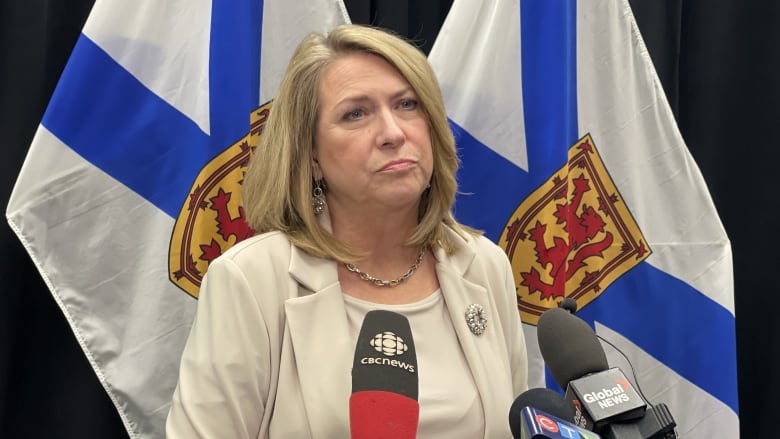 Woman in beige blazer and beige top stands in front of three microphones.