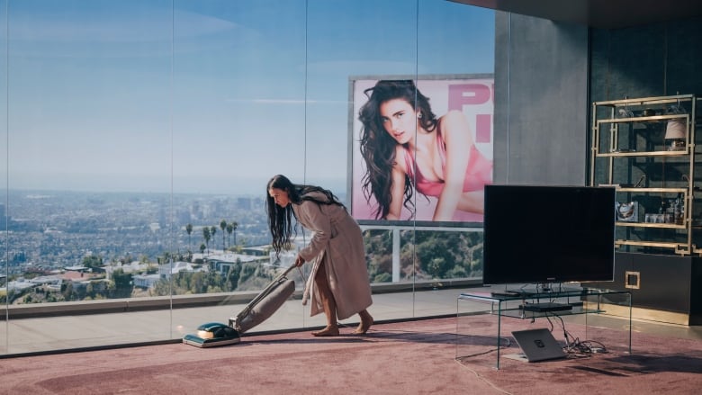 Middle aged woman in luxury home struggles to vacuum while poster of younger woman looks on.