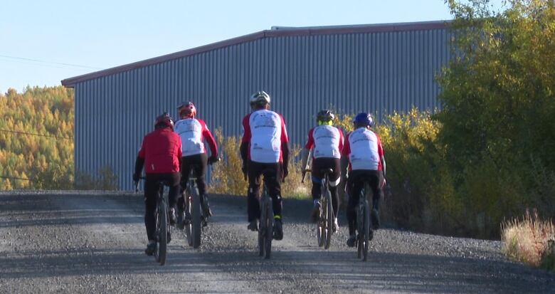 A group of 5 cyclists are seen from the back riding away down a gravel road, in a group. 