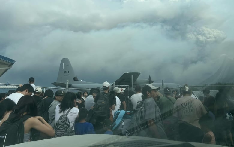 A group of people are grouped behind a fence, on the background there is a military Hercules plane. 