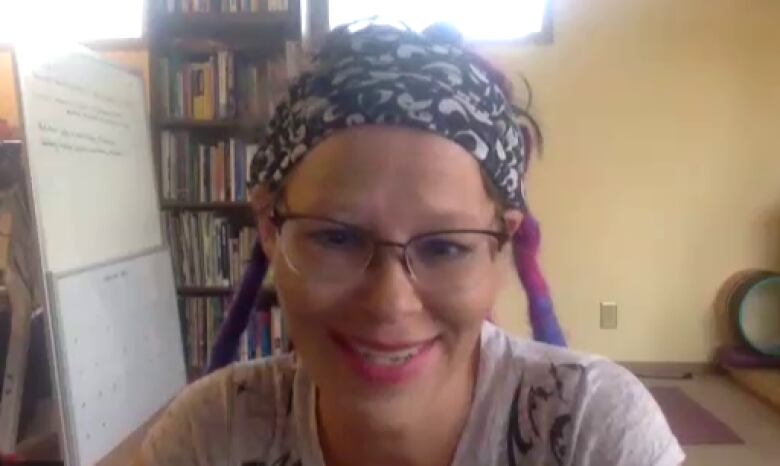 A woman smiles on a Zoom call. Behind her is a yoga mat, a bookshelf and a whiteboard.