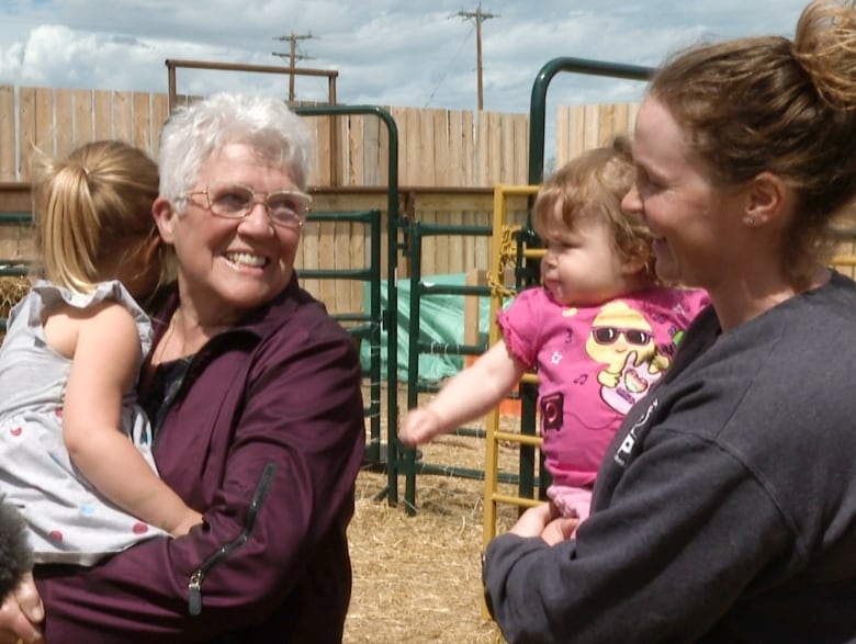 An elderly woman holds a small child and smiles at a younger woman, holding another child. 