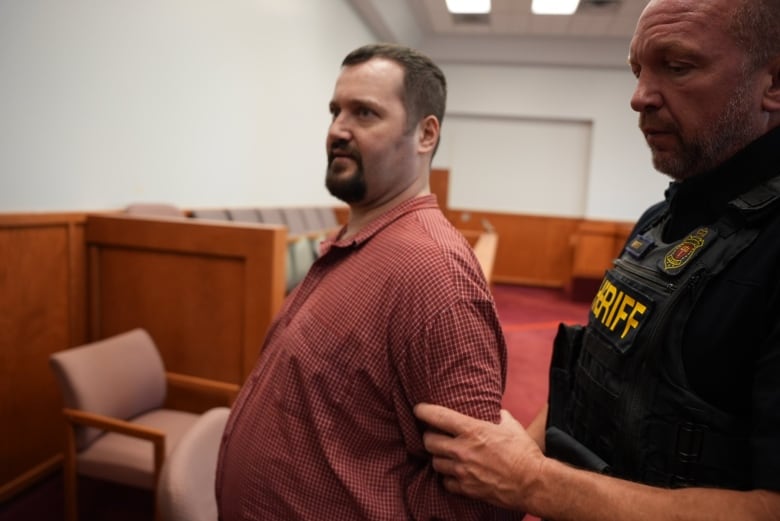 Man in red plaid shirt is held by a sheriff inside court room