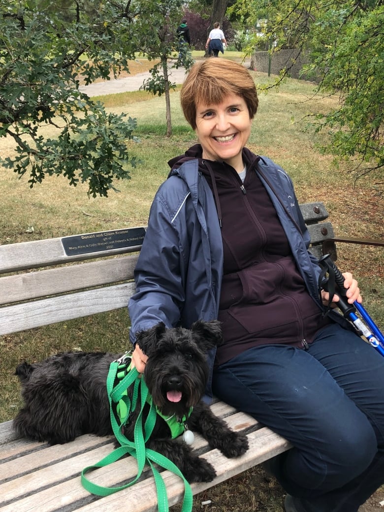 A woman with a short auburn bob sits on a green bench with a little black dog lying beside her.