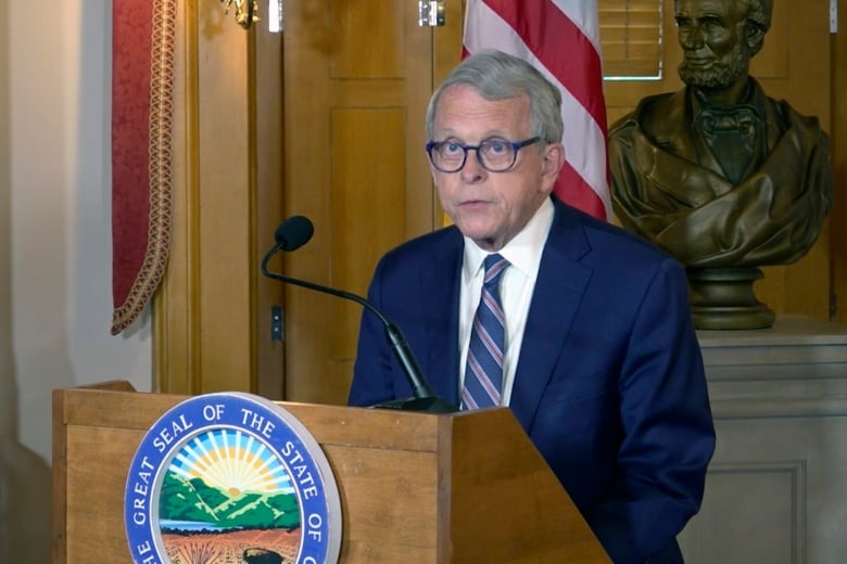 A bespectacled, clean shaven older man is shown speaking at a podium in front of an American flag and a bust of Abraham Lincoln.