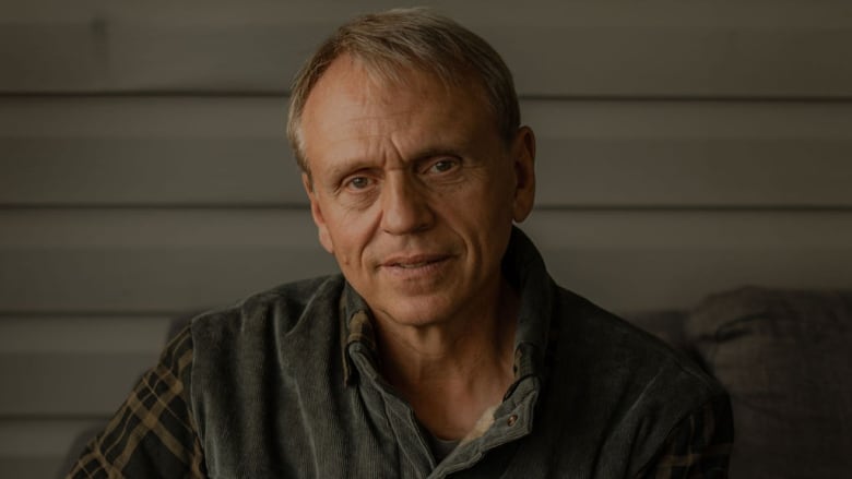 A man in a plaid shirt sits in front of a wood-panelled wall.