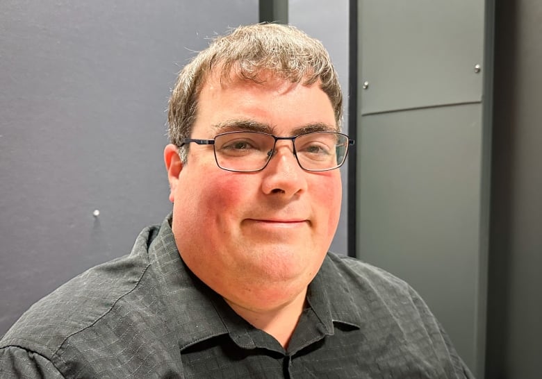 A man wearing a dark grey shirt and glasses sits and smiles in a room with dark grey walls.