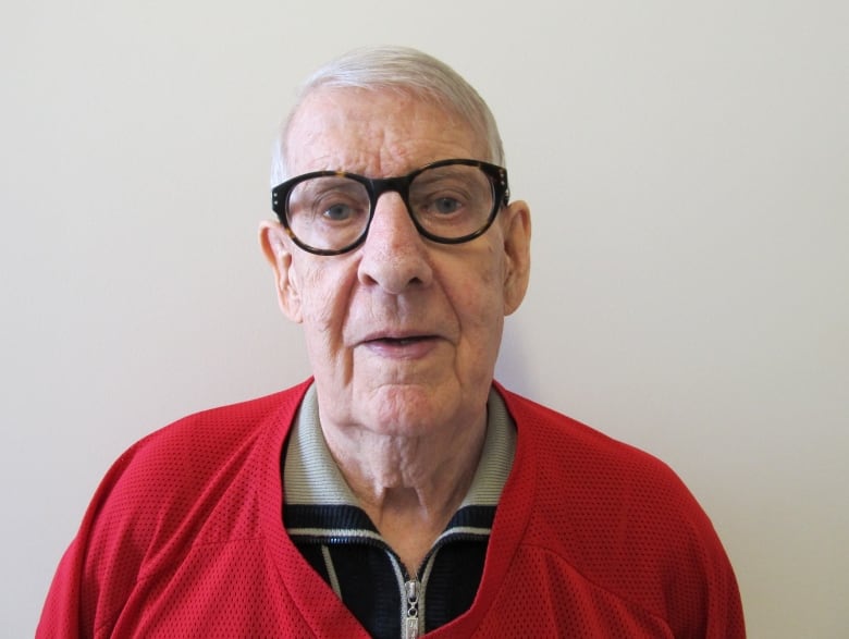 An elderly man with white hair wearing dark-rimmed glasses and a red practice hockey jersey stares into the camera.