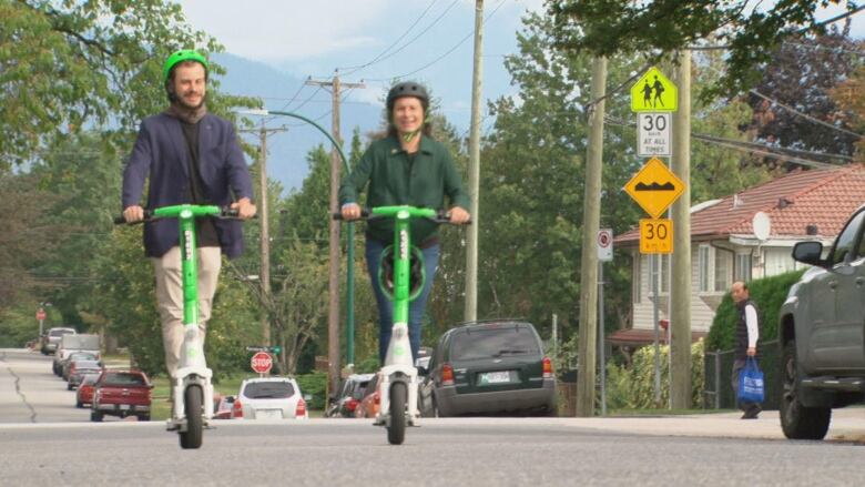 Two people are pictured on a street riding e-scooters. 