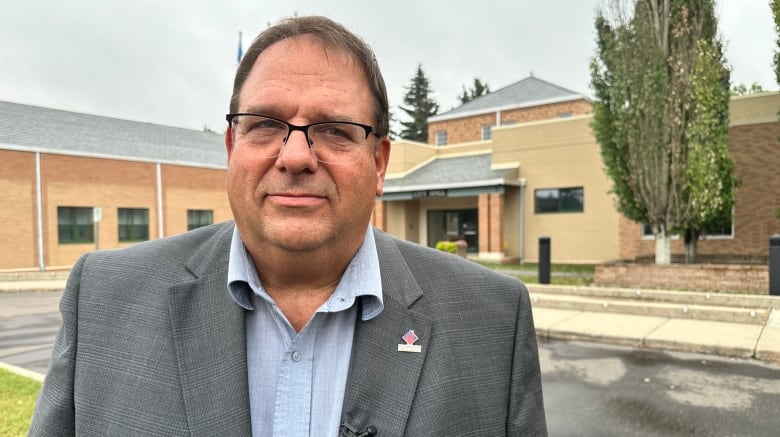 A white man is standing outside, in front of a government building. He is wearing glasses and a blue dress shirt under a grey blazer.