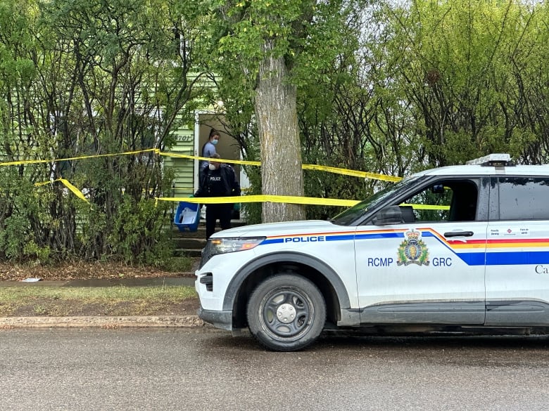 Two investigators exit a green house, fenced in by tall shrubs and trees that have yellow police tape wrapped around the property. A white RCMP cruiser is parked beside the curb.