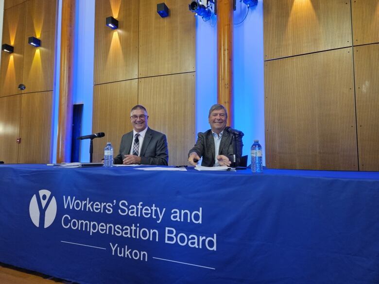 two people sitting at a long table, looking at the camera and smiling, two water bottles on the table, table has a banner that says Workers' Safety and Compensation Board Yukon