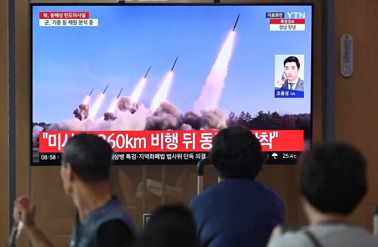 People are shown, backs to the camera, watching a television screen on the wall which shows illustrations of missiles being fired in the sky.