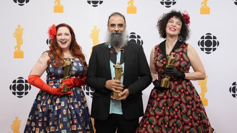 Two happy women in colourful blue and red dresses flank a man in a suit holding a gold trophy.