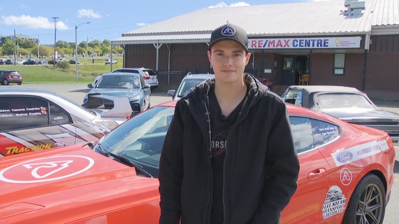 Young man with black baseball hat and clothes stands in front of red sports car smiling.