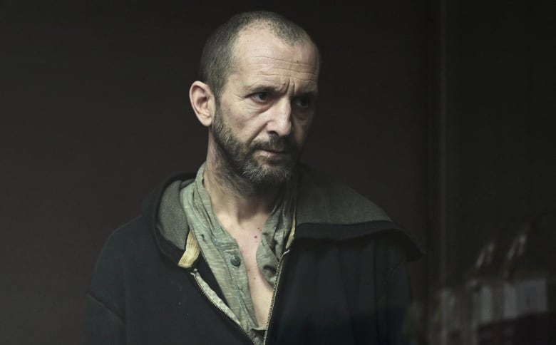 A man with a dark beard stands in a courtroom wearing dark clothing.