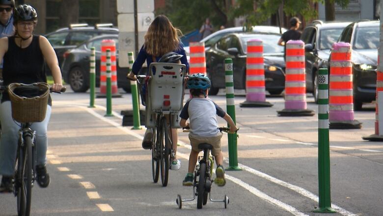 Picture of a bike lane with four bikers riding. Two bikers on the left lane are heading toward the camera. On the right lane, a boy is seen riding a bike with training wheels. He's wearing a blue helmet. He follows behind a woman biking without a helmet. A young child is sitting on a bike seat behind her, wearing a helmet. 