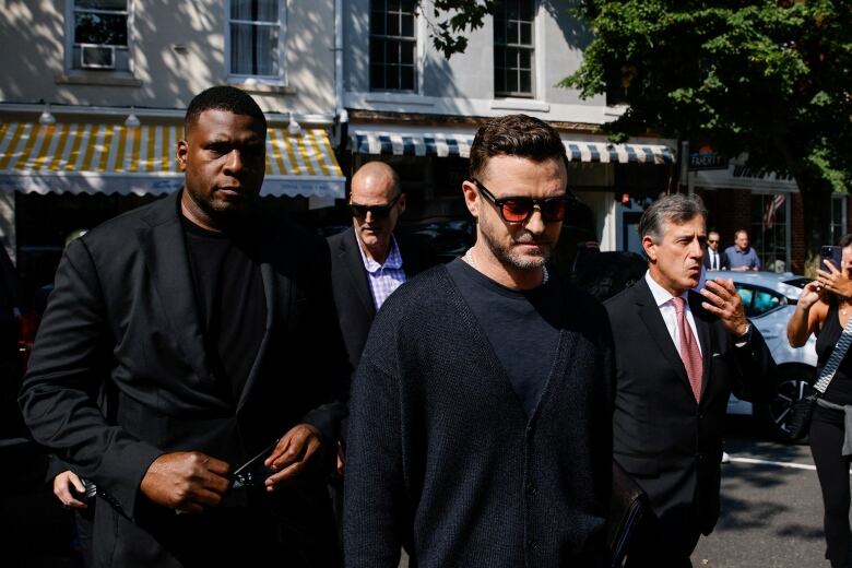 A man in a black sweater and sunglasses with orange lenses walks into court accompanied by a personal security guards.