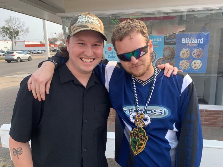 Two men pose together outside a fast food drive-thru on a sunny day.