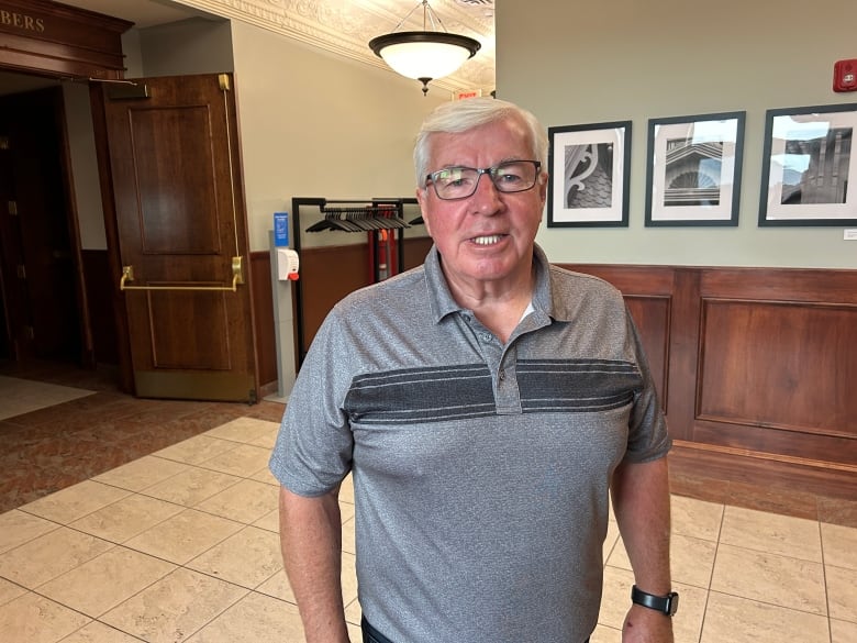 A man with short white hair wearing a polo shirt and glasses standing in a foyer.