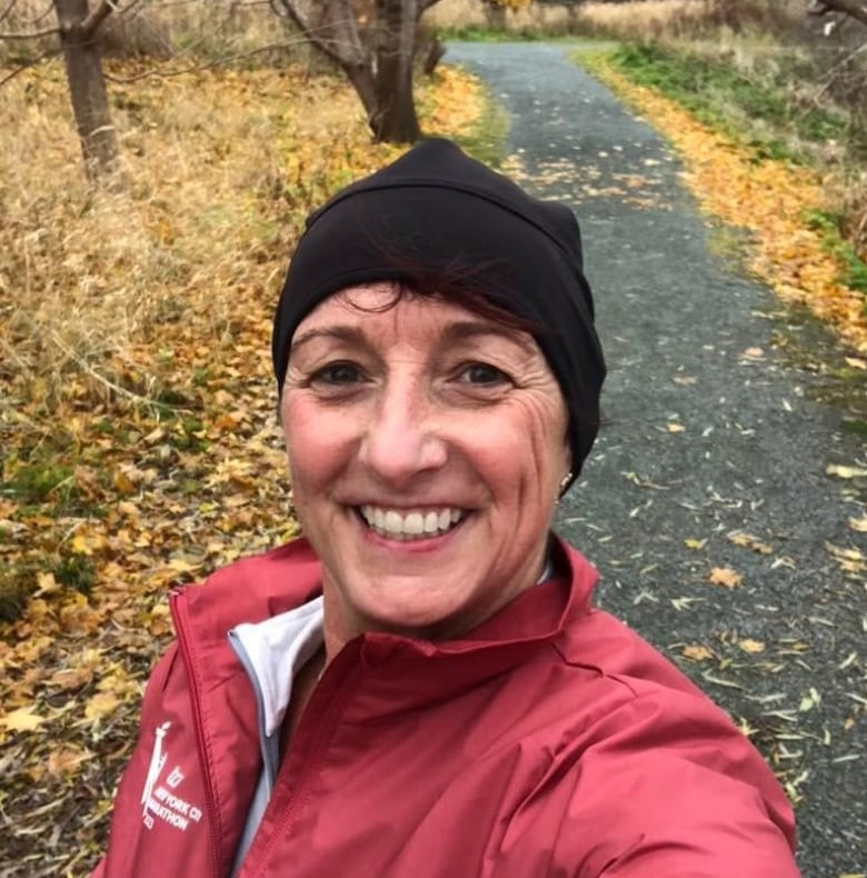 A woman in a red coat and black woolen hat smiles for a selfie. She's standing on a path and there are leaves on the ground.