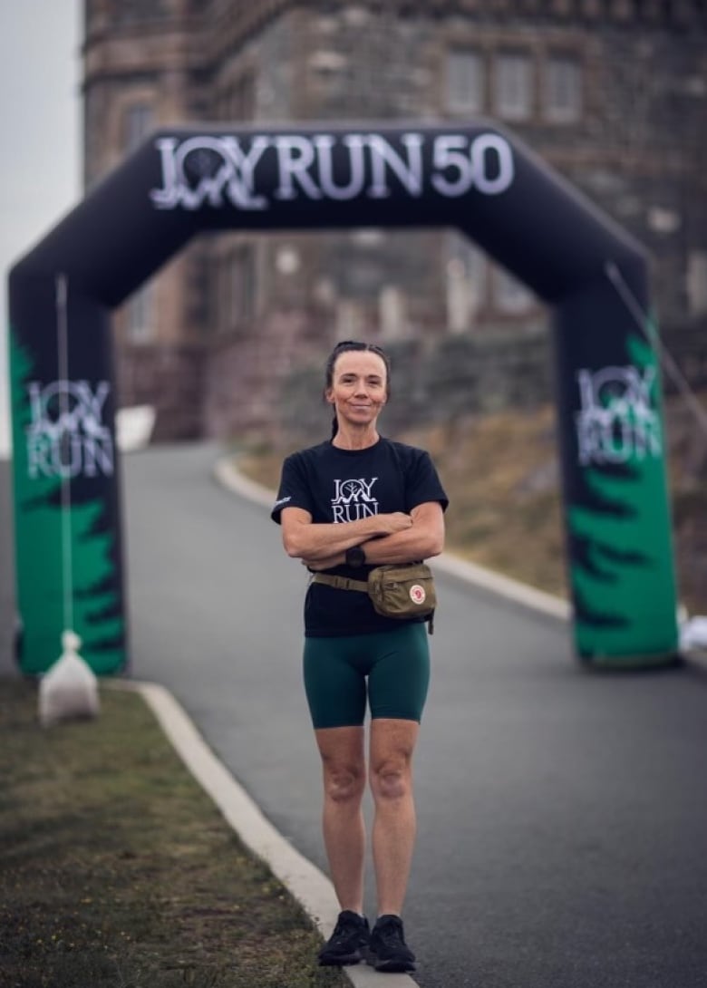 A woman in a black t-shirt and green running shorts stands in front of a black archway that reads Joy Run 50 with her arms folded.