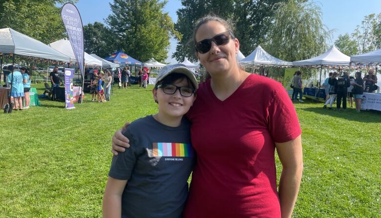 A woman on the right in a red t-shirt and sunglasses hold her son, on the left. He's wearing a tan cap, glasses and a black shirt with a Pride motif. 