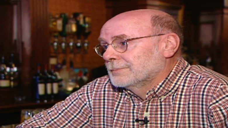 A bearded man wearing glasses and a dress shirt is shown inside of a bar.