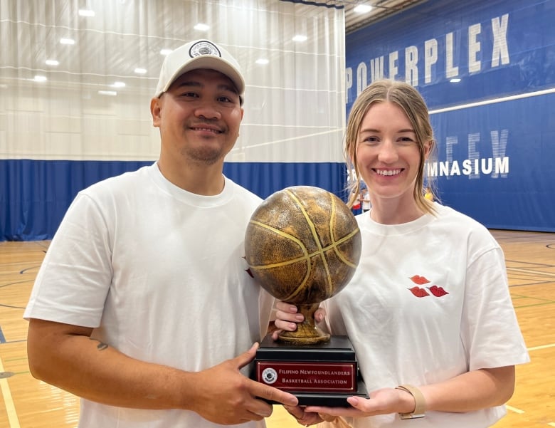 A man, left, and a woman, right, smile. Together, they hold a trophy in the shape of a basketball. They stand on a basketball court.