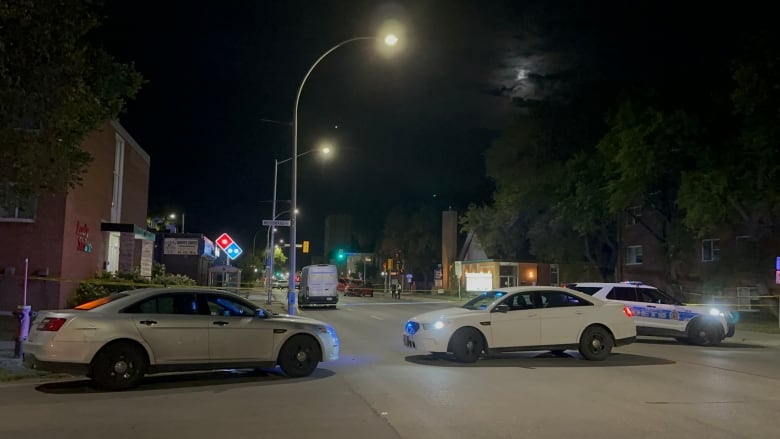 One police vehicle and two unmarked police cars block off a section of a street where a red truck appears severely damaged. 