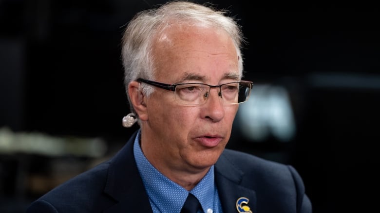 A white man wearing a blue suit with a 'C' pin on it is pictured talking.