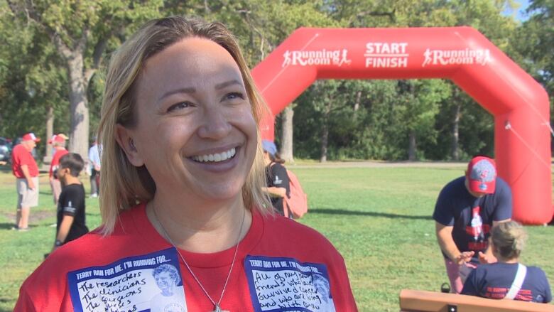 A woman wearing a red shirt with writing on it smiles while talking to a reporter outside. 