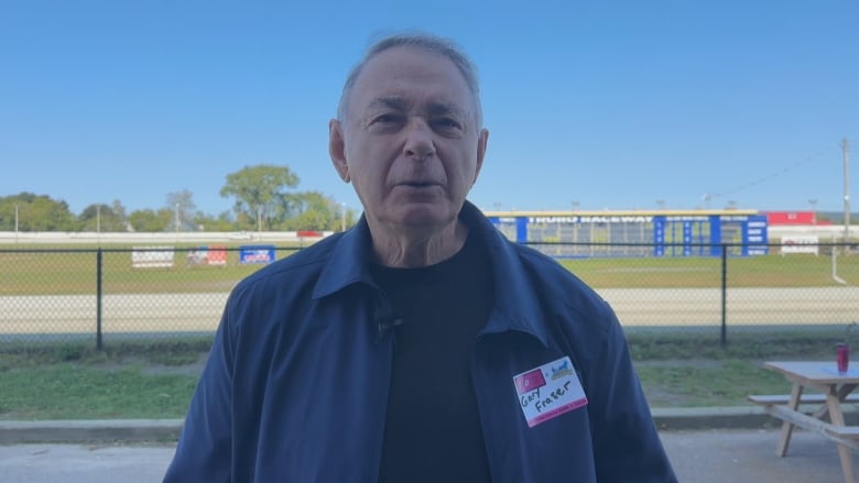 A man in a blue jacket and t-shirt stands in front of a horseracing track.