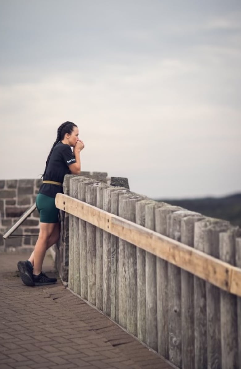 A woman in a black t-shirt and green running shorts is leaning against a railing with her hands clasped over her mouth. She is deep in thought.