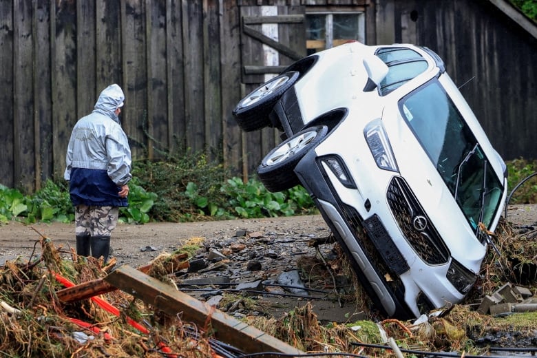 A person in rain gear stands near an SUV up on two wheels.