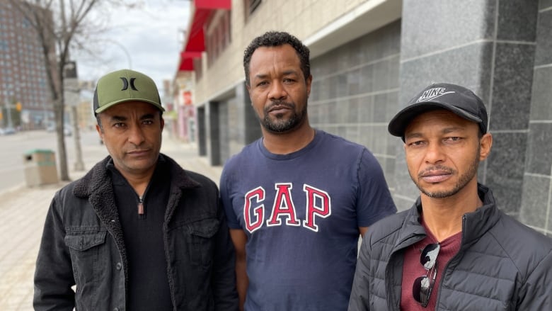 A portrait of three men outside; the first man is wearing a black shirt and dark jeans, the second man is wearing a t-shirt and blue jeans and the third man is wearing a ball cap and a jacket.