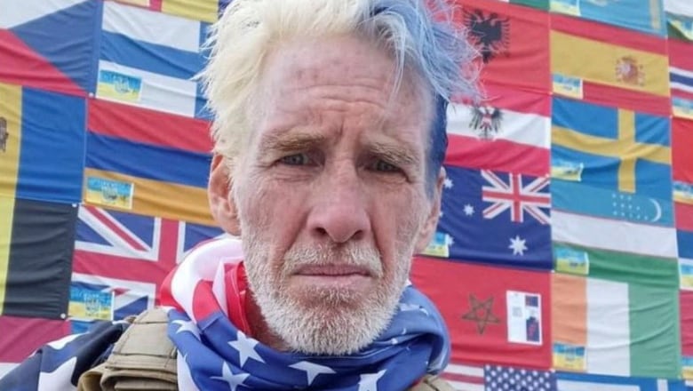 A man with white hair and a white beard is seen wearing a combat vest and bandana printed with the American flag around his neck.