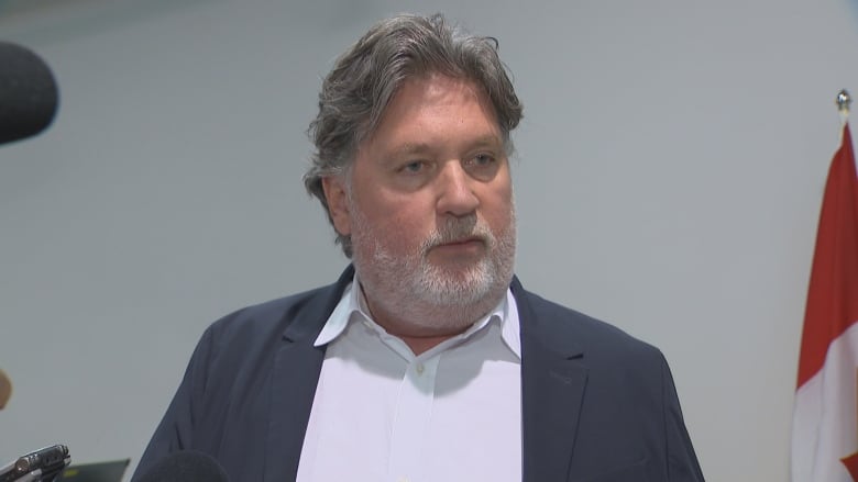 Man in blazer and white shirt stands near a red and white flag.