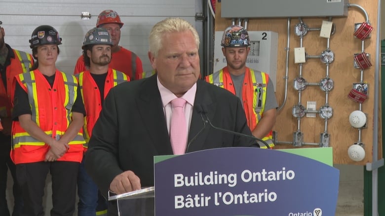 Doug Ford standing at a podium with workers in hard hats and hi-viz vests lined up  behind him.