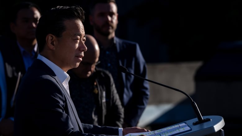 A man is pictured in profile speaking at a podium, with other people in the background.